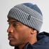 Man wearing a gray and blue knit beanie with a brand logo on a light gray background